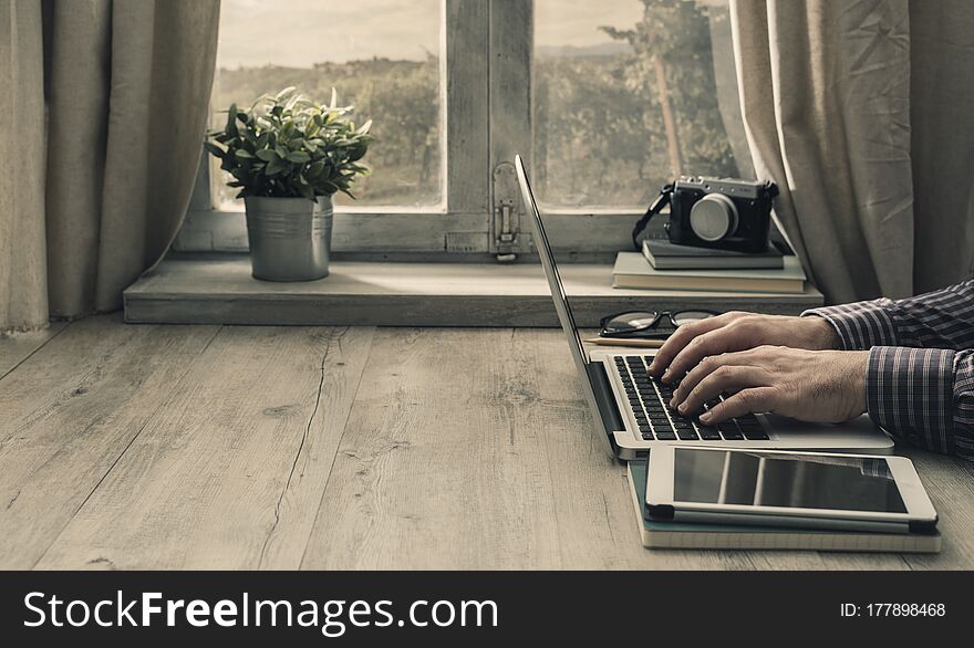 Hipster man working with a laptop and a tablet in front of a window in a rustic vintage house. Hipster man working with a laptop and a tablet in front of a window in a rustic vintage house