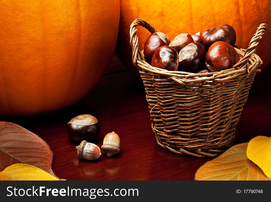 Two pumpkins, colored leaves, acorns, and a basket of chestnuts on a warm wooden tabletop. Two pumpkins, colored leaves, acorns, and a basket of chestnuts on a warm wooden tabletop.