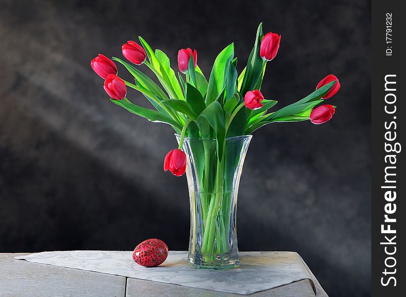 A vase of red tulips on a tiled table with a decorated egg and sunbeams in the background. A vase of red tulips on a tiled table with a decorated egg and sunbeams in the background.