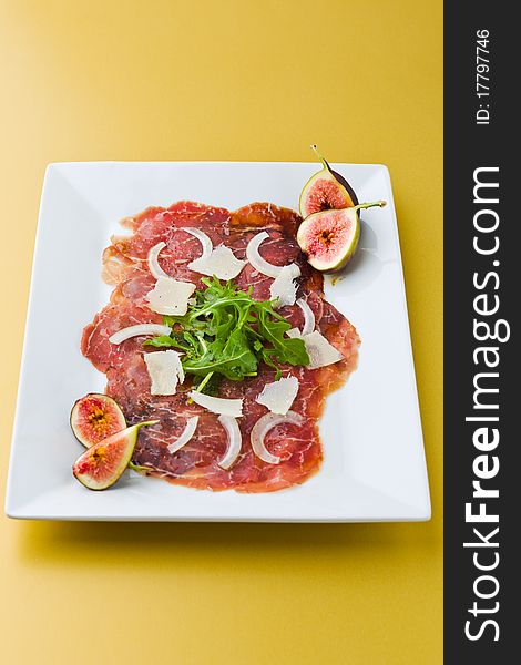 Red beef carpaccio in a white plate with salad, fig fruit, parmesan cheese and olive oil. Yellow background and shallow depth of field. Red beef carpaccio in a white plate with salad, fig fruit, parmesan cheese and olive oil. Yellow background and shallow depth of field.