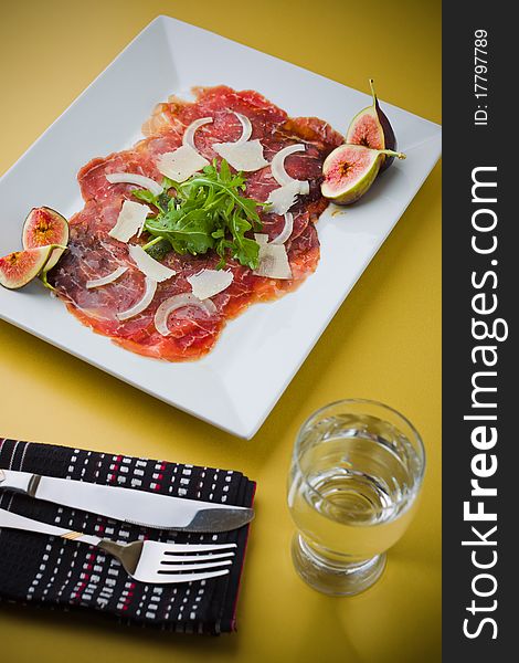 Red beef carpaccio in a white plate with salad, fig fruit, parmesan cheese and olive oil. Yellow background and shallow depth of field. Red beef carpaccio in a white plate with salad, fig fruit, parmesan cheese and olive oil. Yellow background and shallow depth of field.