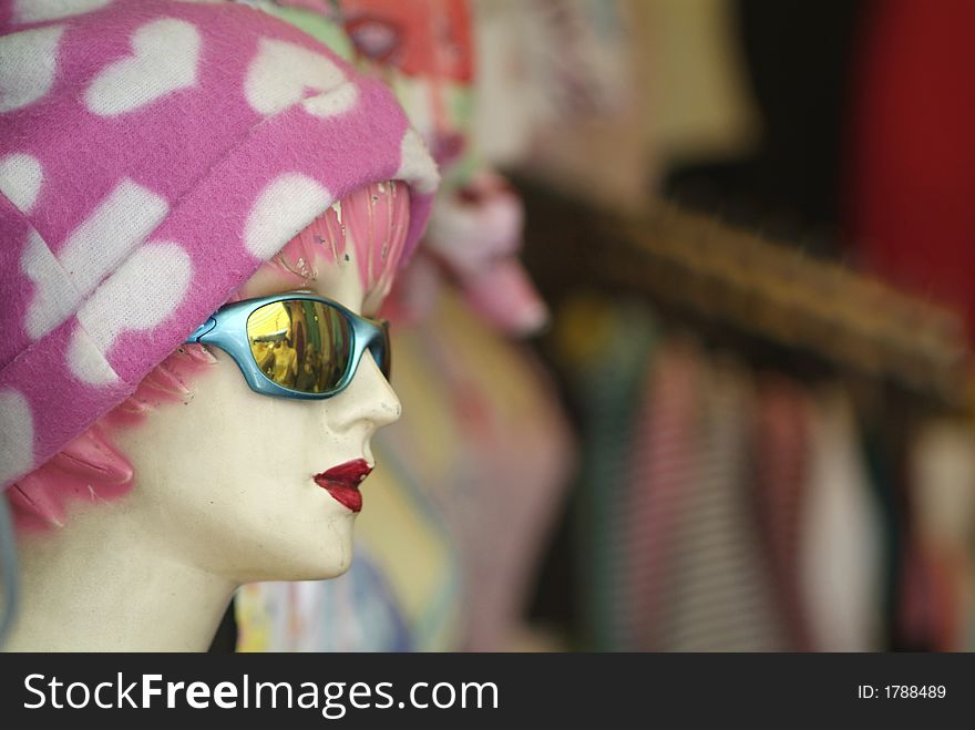 Old, worn mannequin with pink and white cap, sunglasses and deep red lipstick. Blurry, out-of-focus shop-background in the right two thirds of the photo. Old, worn mannequin with pink and white cap, sunglasses and deep red lipstick. Blurry, out-of-focus shop-background in the right two thirds of the photo.
