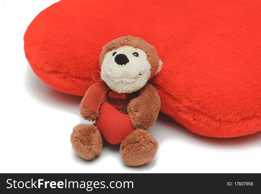 Little teddy-bear holding red heart. Bigger red pillow-heart behind it. White isolated background