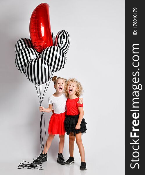 Two happy singing screaming blonde kids girls in red and black skirts and white and red t-shirts with big stylish air balloons stand together hugging. Two happy singing screaming blonde kids girls in red and black skirts and white and red t-shirts with big stylish air balloons stand together hugging
