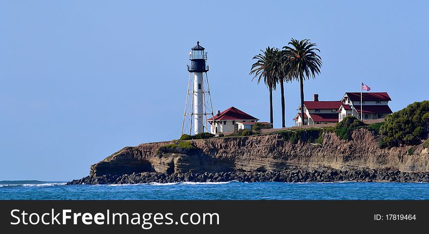 Lighthouse on California coast with buildings, palms, and American flag.