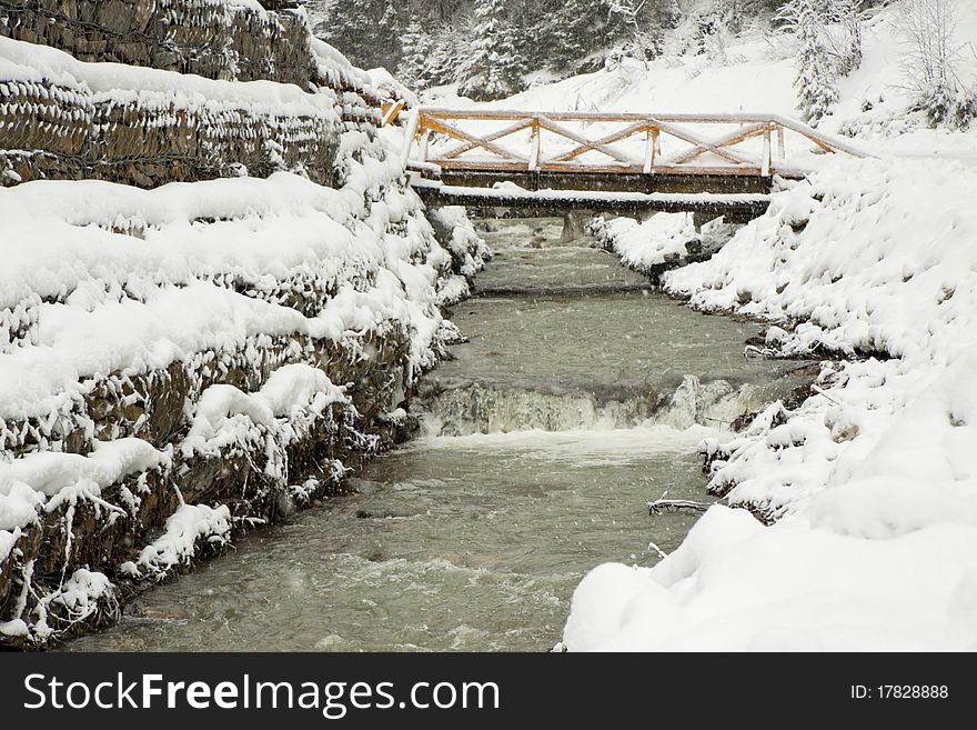 The bridge through the mountain river near wood, under going snow. The bridge through the mountain river near wood, under going snow