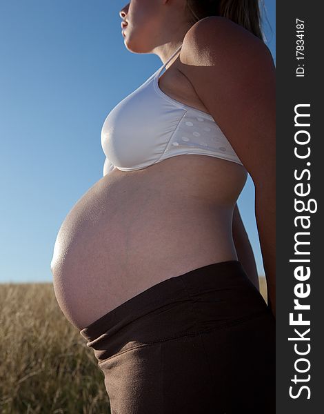 A young pregnant woman standing in an open field wearing a bra and looking peaceful.