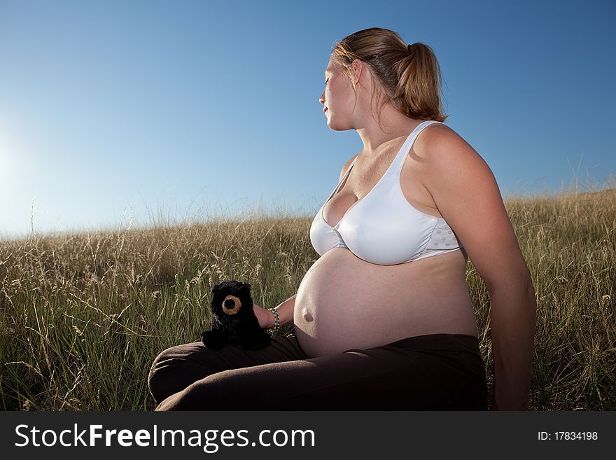 A young pregnant woman sitting in an open field wearing a bra and holding a teddy bear looking very relaxed and peaceful.