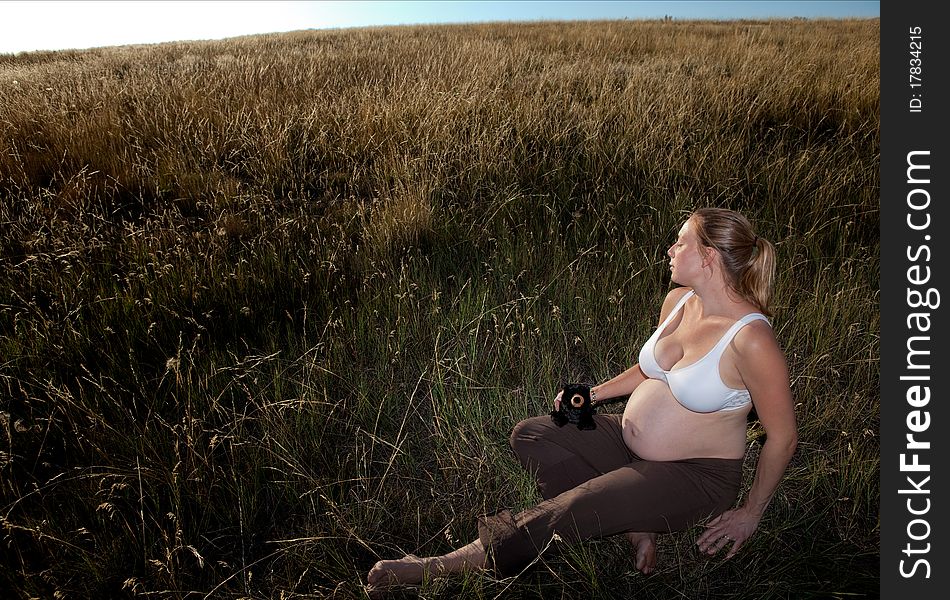 A young pregnant woman sitting in an open field wearing a bra and holding a teddy bear looking very relaxed and peaceful.