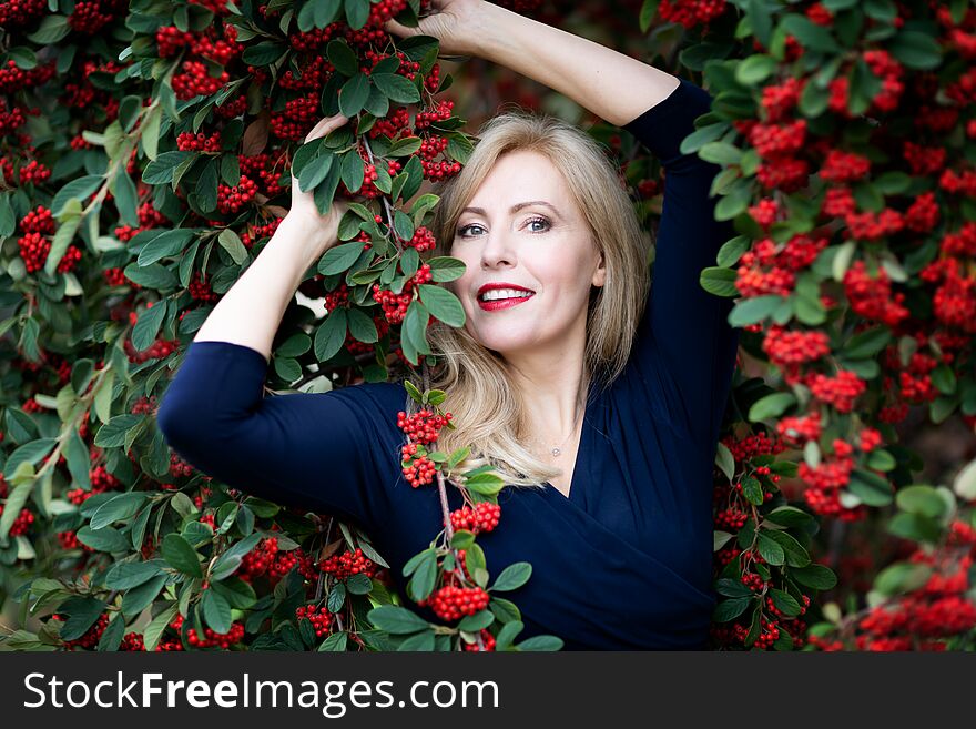 A waist up portrait of Caucasian woman with long blond hair with red near a bush with red berries.