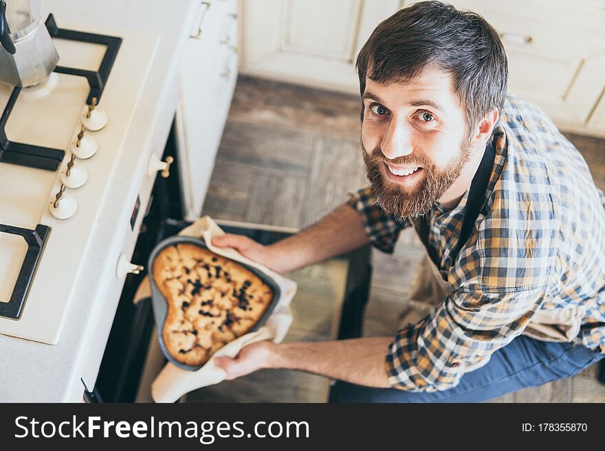 Bearded young man taking heart shaped berry pie out of the oven. Cooking at home and smiling. Bearded young man taking heart shaped berry pie out of the oven. Cooking at home and smiling