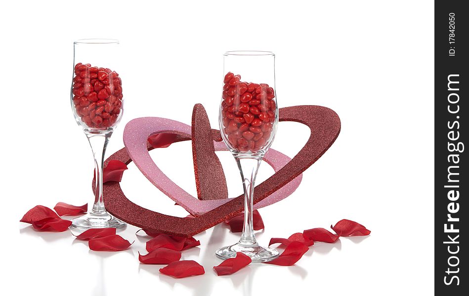 A front closeup view of two champagne glasses filled with cinnamon hearts, with multi-sized glitter valentines and rose petals on the white background. A front closeup view of two champagne glasses filled with cinnamon hearts, with multi-sized glitter valentines and rose petals on the white background.