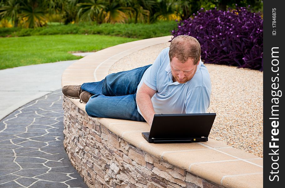 Man lying down on a wall in a park using a laptop computer. Man lying down on a wall in a park using a laptop computer.
