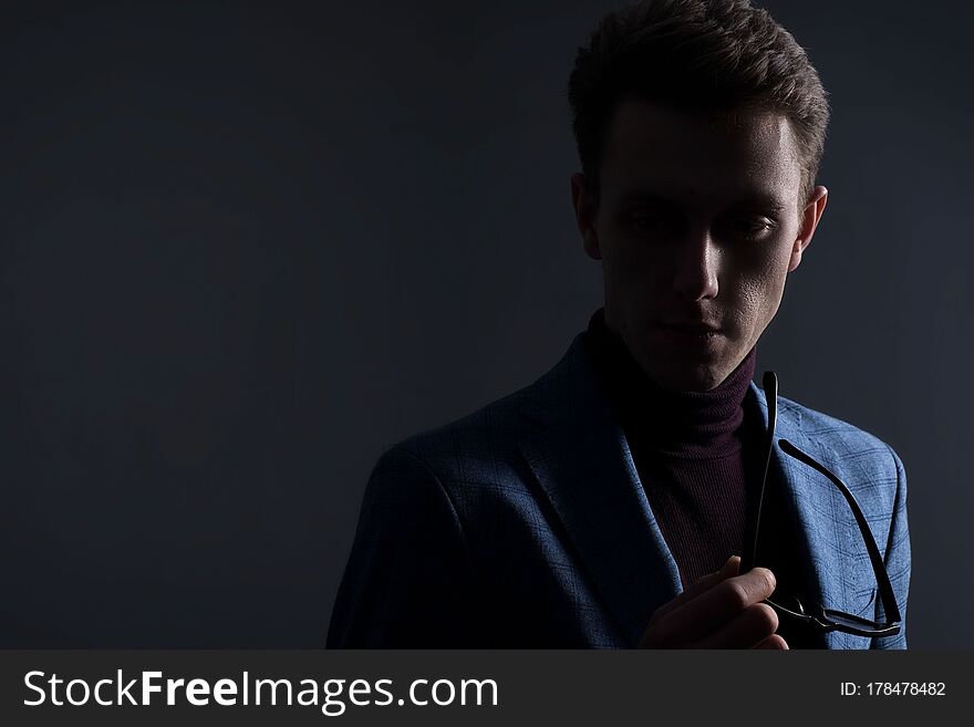 Close-up dramatic portrait in dark colors of a young guy, a businessman in a business suit, looks away, on a gray background. Minimalist portrait. Dark male business strict portrait. Young businessmen. Free space for advertising. Men`s jacket, tie, pants, tuxedo