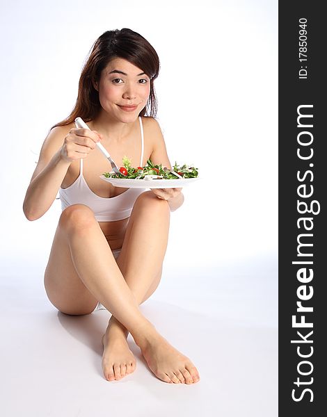 Beautiful young oriental girl, sitting on floor wearing sports underwear, showing off a healthy body while eating a salad. Beautiful young oriental girl, sitting on floor wearing sports underwear, showing off a healthy body while eating a salad.