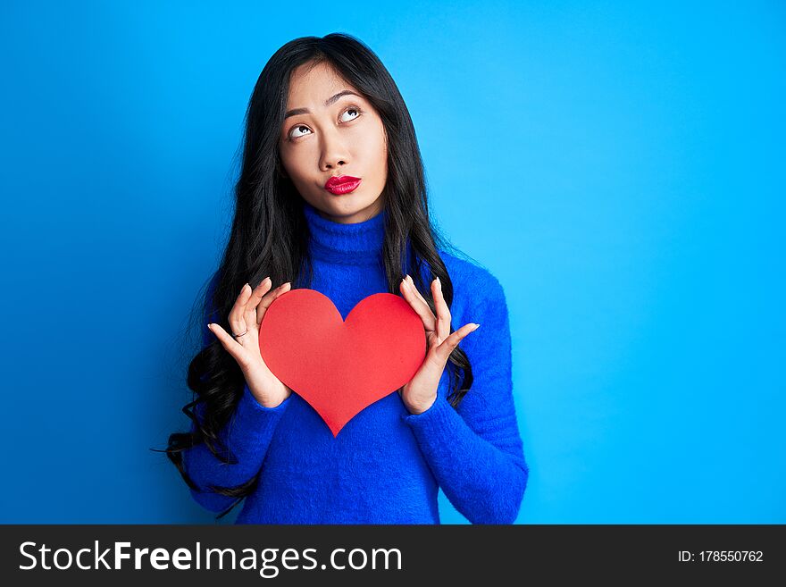 Pretty young woman holding big paper heart in her hands and looking up isolated on blue background. Love and romantic concept