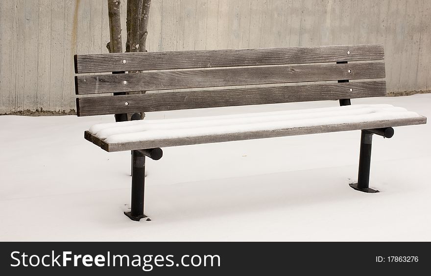 Isolated wooden bench covered with snow sits starkly against cement wall. Isolated wooden bench covered with snow sits starkly against cement wall.