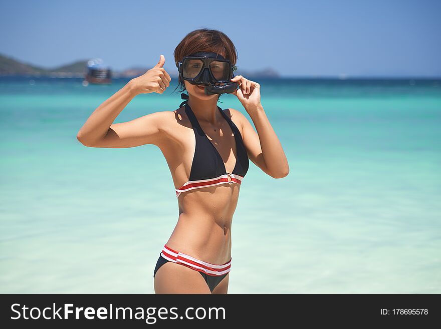 Beautiful young girl with slim build tanned skin, mask and breathing tube, short Bob haircut wears black swimsuit with red border against background of turquoise sea, blue sky and rolling mountains. Beautiful young girl with slim build tanned skin, mask and breathing tube, short Bob haircut wears black swimsuit with red border against background of turquoise sea, blue sky and rolling mountains.