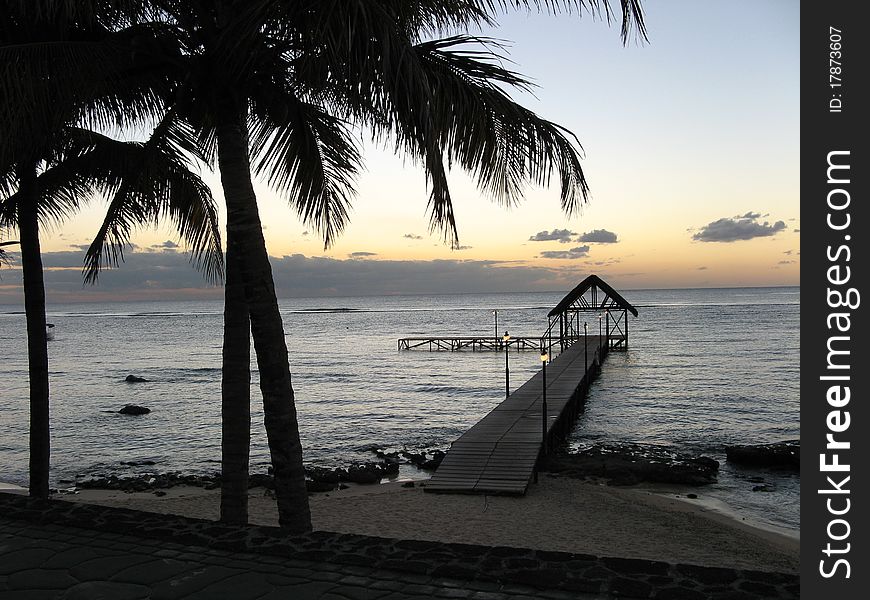 Silhouette of a tropical beach house, jetty and palm trees at sunset.