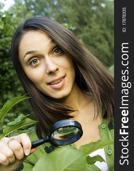 Young woman outdoors looking through a magnifying glass on the green leaves of the tree. Young woman outdoors looking through a magnifying glass on the green leaves of the tree