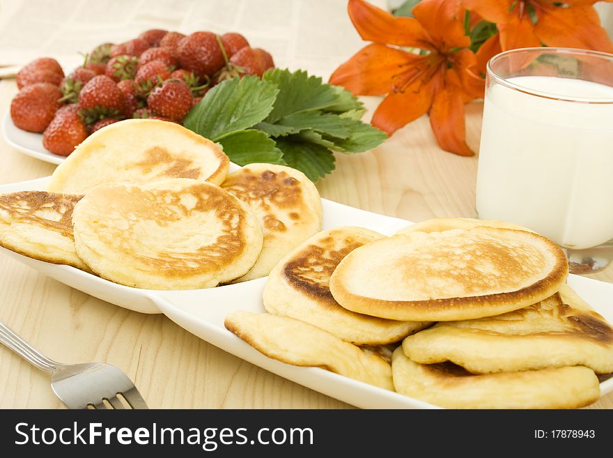 On the table a plate of pancakes, a plate of strawberries, a glass of milk and orange lilies. Breakfast