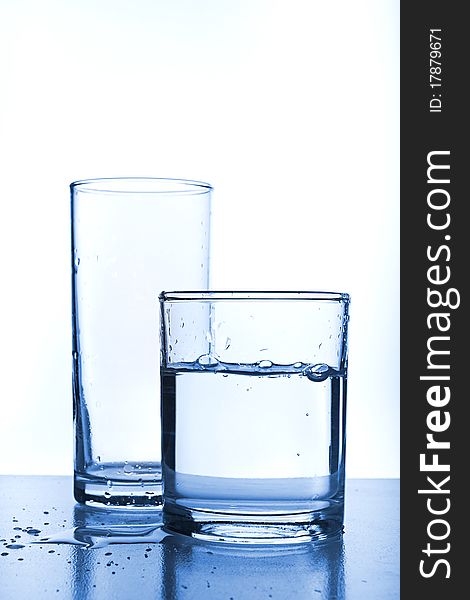 Empty glass behind full glass of water. Image stylized in blue tone for more cool feeling. Empty glass behind full glass of water. Image stylized in blue tone for more cool feeling