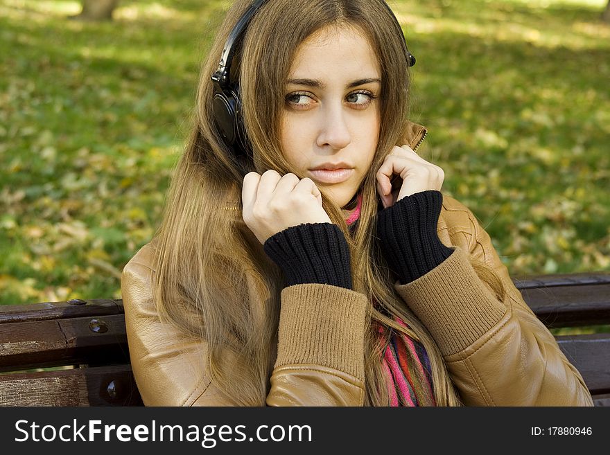 Young Caucasian woman with headphones in autumn park sitting on a wooden bench. Autumn around a lot of colorful foliage. Young Caucasian woman with headphones in autumn park sitting on a wooden bench. Autumn around a lot of colorful foliage