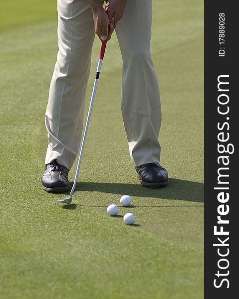 A golfer wearing beige pants and black leather shoes practicing his putt with several balls on a sunny day. A golfer wearing beige pants and black leather shoes practicing his putt with several balls on a sunny day