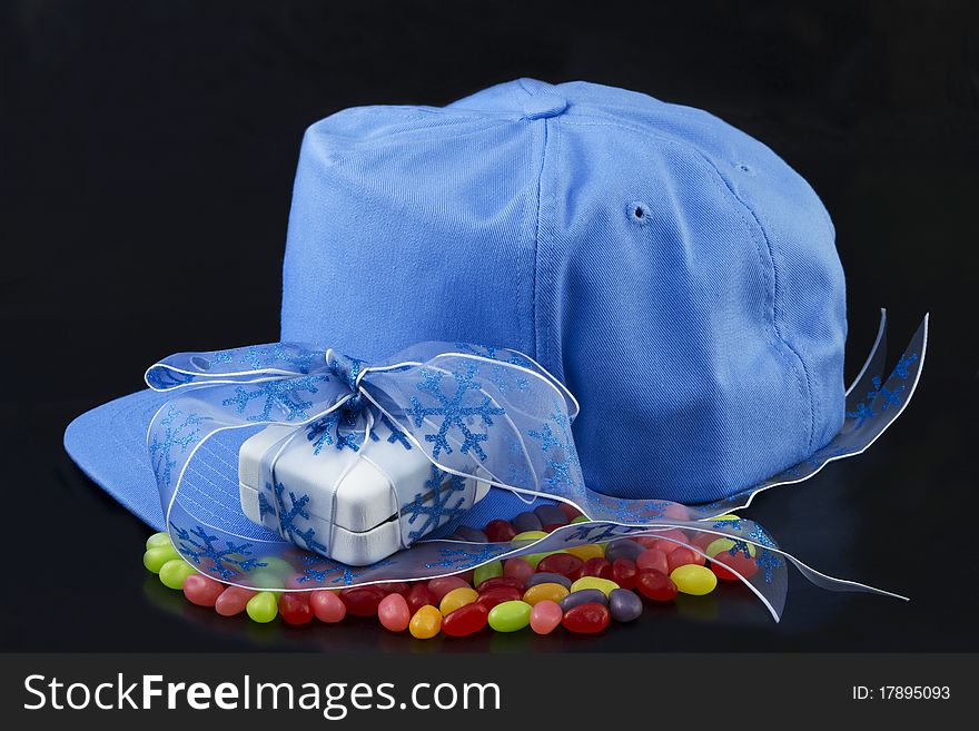 Blue baseball cap, blue ribbon around a white jewelry box placed on colorful jellybeans on black background reflect the sweet romance of love and sports. Blue baseball cap, blue ribbon around a white jewelry box placed on colorful jellybeans on black background reflect the sweet romance of love and sports