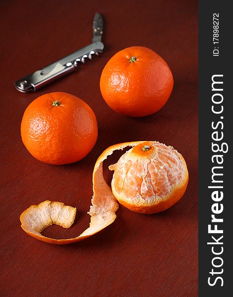 Three Mandarin Oranges, one of which is peeled, on a dark wood background with a pen knife