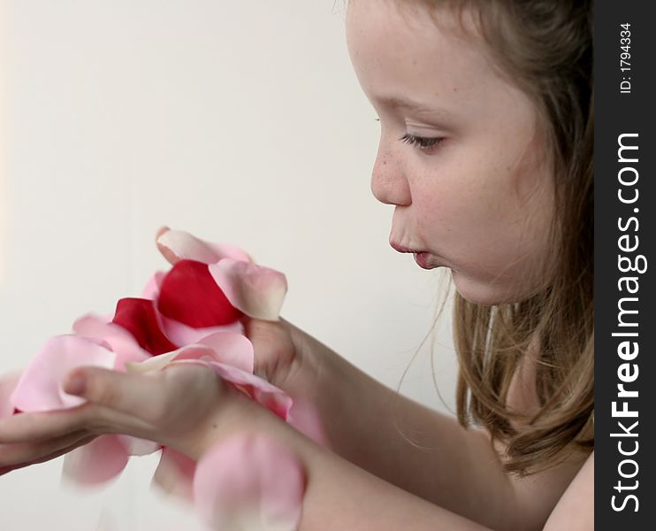 Young girl blowing pink, red, white rose petals held in hands over a slightly pink background. Young girl blowing pink, red, white rose petals held in hands over a slightly pink background