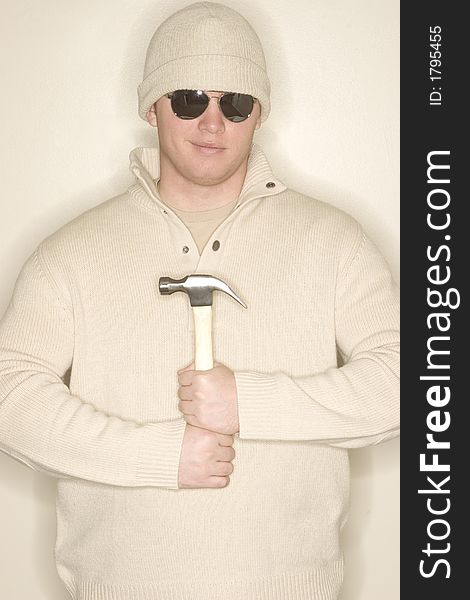 Man smiles as he holds a hammer to his chest as he wears sunglasses, a wool hat, and a wool sweater. Man smiles as he holds a hammer to his chest as he wears sunglasses, a wool hat, and a wool sweater