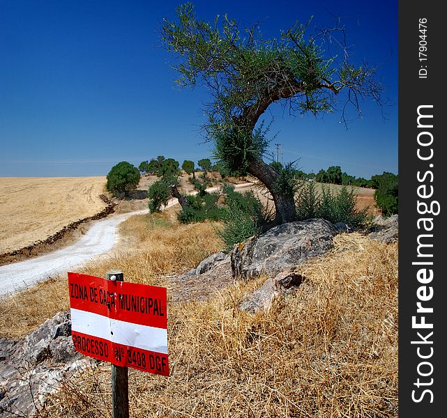 White and red sign with a country road under blue sky. Trees on fields with grass burned by the sun. White and red sign with a country road under blue sky. Trees on fields with grass burned by the sun