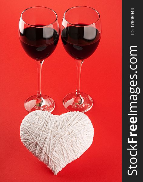 Two glasses of wine and a white heart made of wool on a red background. Valentine's Day