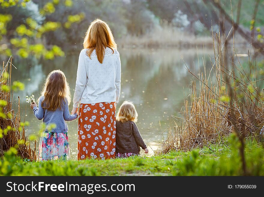 Redhead mom with two daughters standing on the banks of a small pond. spring landscape near the water. rear view. Redhead mom with two daughters standing on the banks of a small pond. spring landscape near the water. rear view.