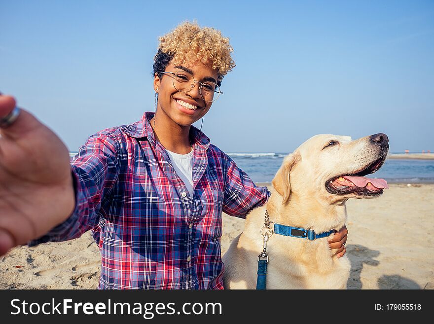 Happy african american woman hugging her dog in tropical beach. she makes selfie on camera smart phone .