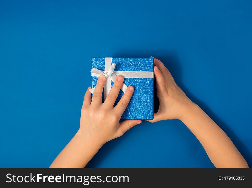 Children`s hands hold a blue shiny box with Christmas with a gift on a blue background