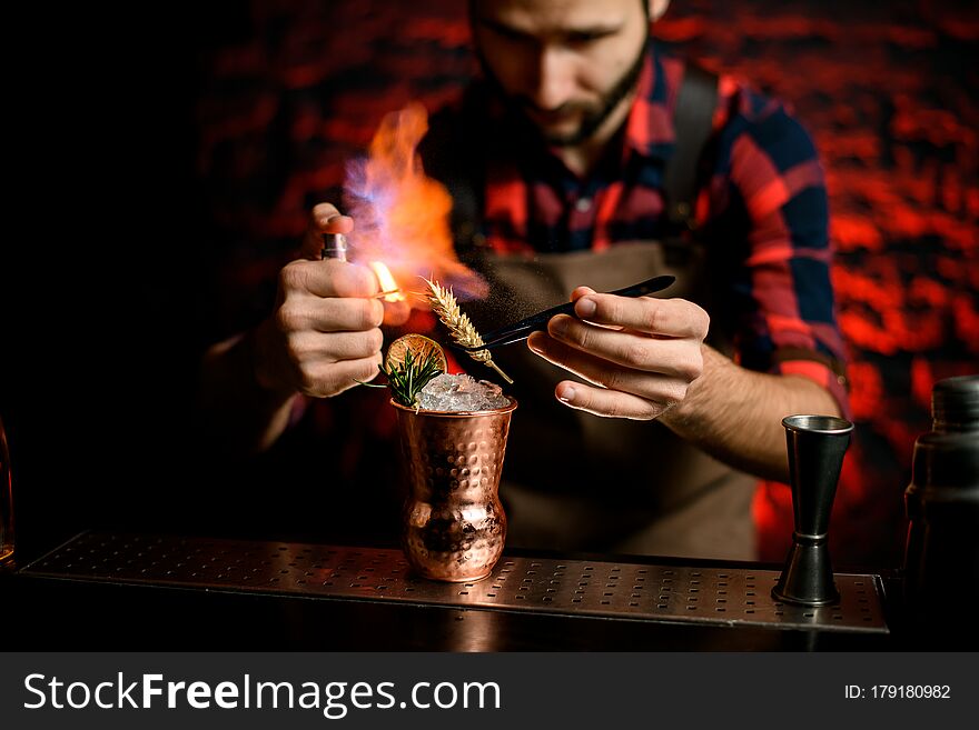 Bartender holds spikelet using tweezers over metal glass with cold cocktail spray and makes fire. Bartender holds spikelet using tweezers over metal glass with cold cocktail spray and makes fire.