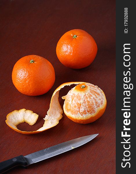 Three Mandarin Oranges, one of which is peeled, on a dark wood background with a kitchen knife
