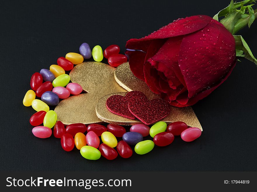 A single, red rose is placed on a black background with wo gold hears, two red hearts, and colorful jelly beans;. A single, red rose is placed on a black background with wo gold hears, two red hearts, and colorful jelly beans;