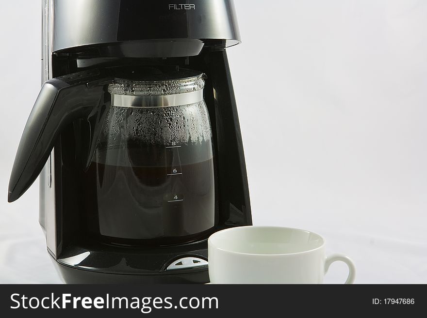 A black coffee machine with a glass jug full of hot freshly made coffee and a white cup waiting to be filled up. A black coffee machine with a glass jug full of hot freshly made coffee and a white cup waiting to be filled up