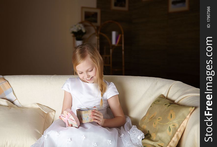 A beautiful little girl in white dress sitting on a sofa with a present at home. A beautiful little girl in white dress sitting on a sofa with a present at home