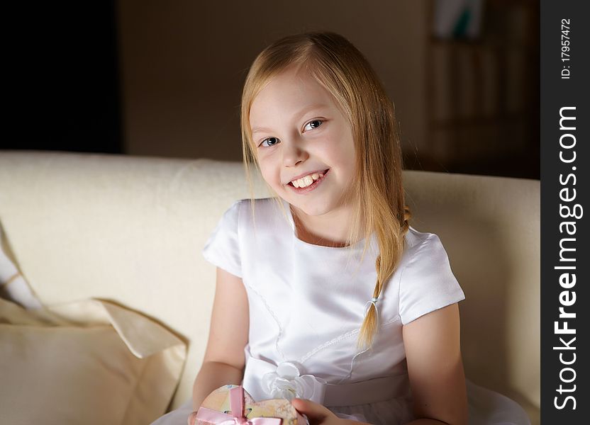 A beautiful little girl in white dress sitting on a sofa with a present at home. A beautiful little girl in white dress sitting on a sofa with a present at home