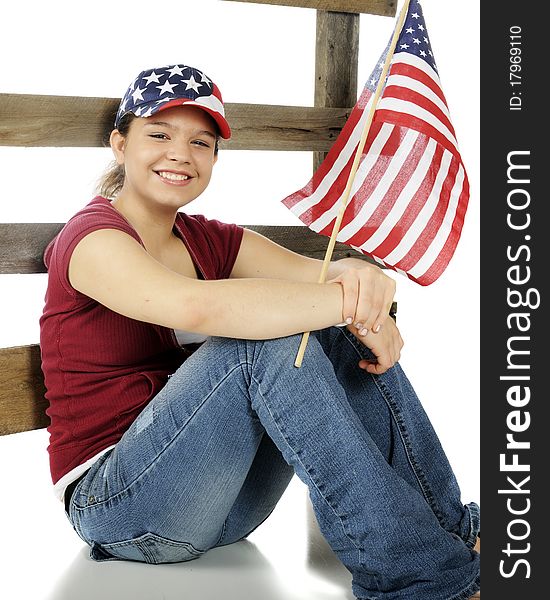 A pretty young teen sitting against a fence, wearing a stars-and-stripes hat and holding an American flag. Isolated on white. A pretty young teen sitting against a fence, wearing a stars-and-stripes hat and holding an American flag. Isolated on white.