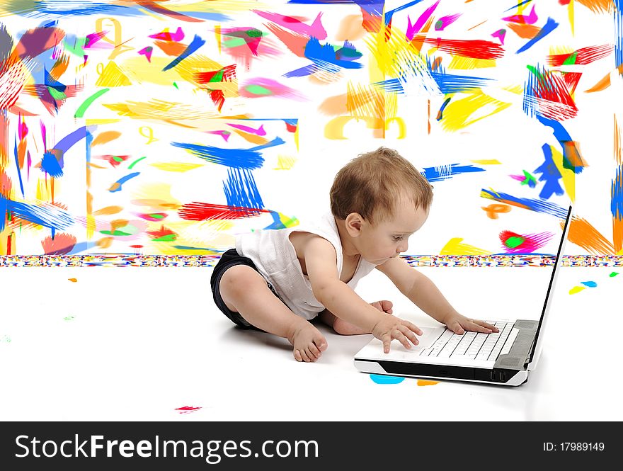 Little baby boy is sitting on floor with his laptop, over white wall, in messy painted room with many colors around