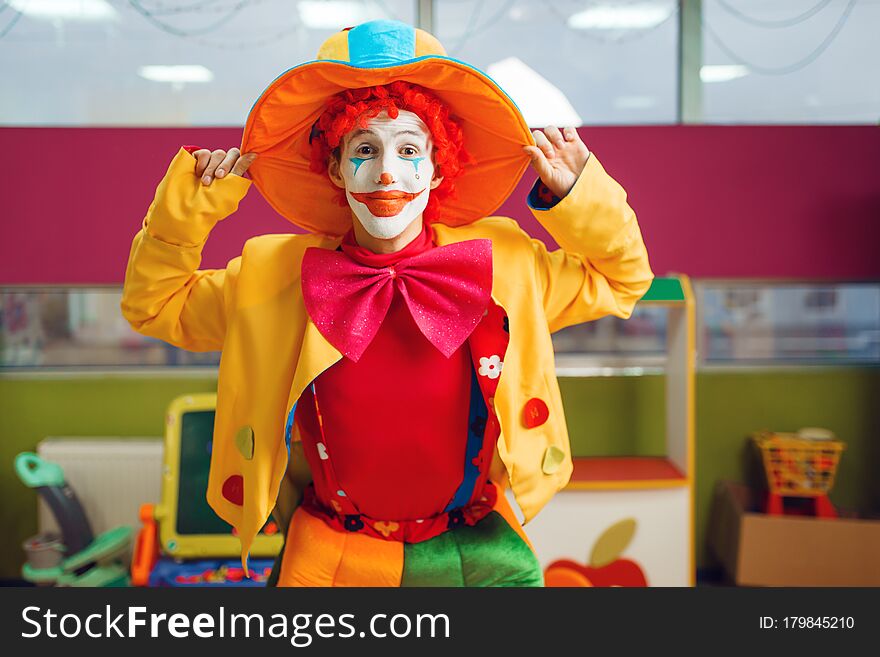Funny clown with makeup dressed in colorful hat and costume poses in children`s area. Birthday party in playroom, baby holiday in playground. Childish leisure, entertainment with animator. Funny clown with makeup dressed in colorful hat and costume poses in children`s area. Birthday party in playroom, baby holiday in playground. Childish leisure, entertainment with animator