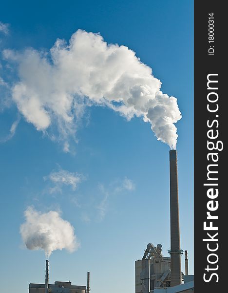 A manufacturing facility pumps smoke into the air from two smokestacks with a clear blue sky in the background. A manufacturing facility pumps smoke into the air from two smokestacks with a clear blue sky in the background.