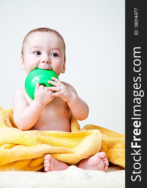 Baby boy after bath wrapped in soft and fluffy yellow towel sitting holding and gnawing a green circular toy with his feet peeking from the towel and smiling. Baby boy after bath wrapped in soft and fluffy yellow towel sitting holding and gnawing a green circular toy with his feet peeking from the towel and smiling