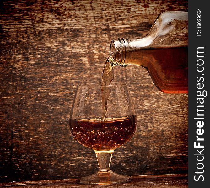 Cognac pour from a bottle in a glass on wood background