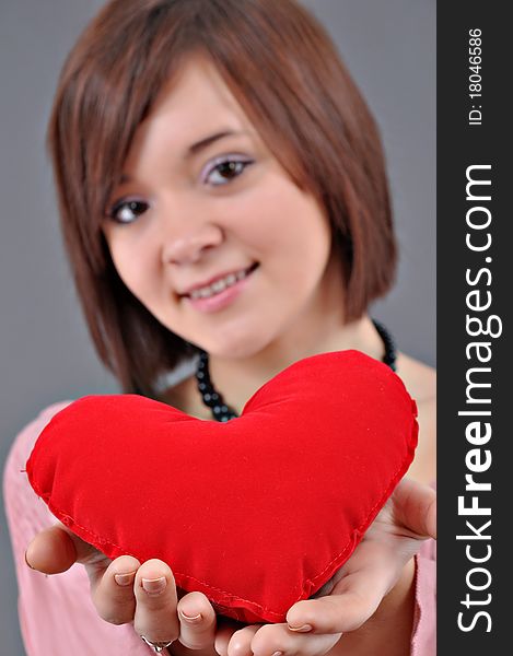 The beautiful young woman holds in hands a red heart on a grey background. Selective focus on heart.